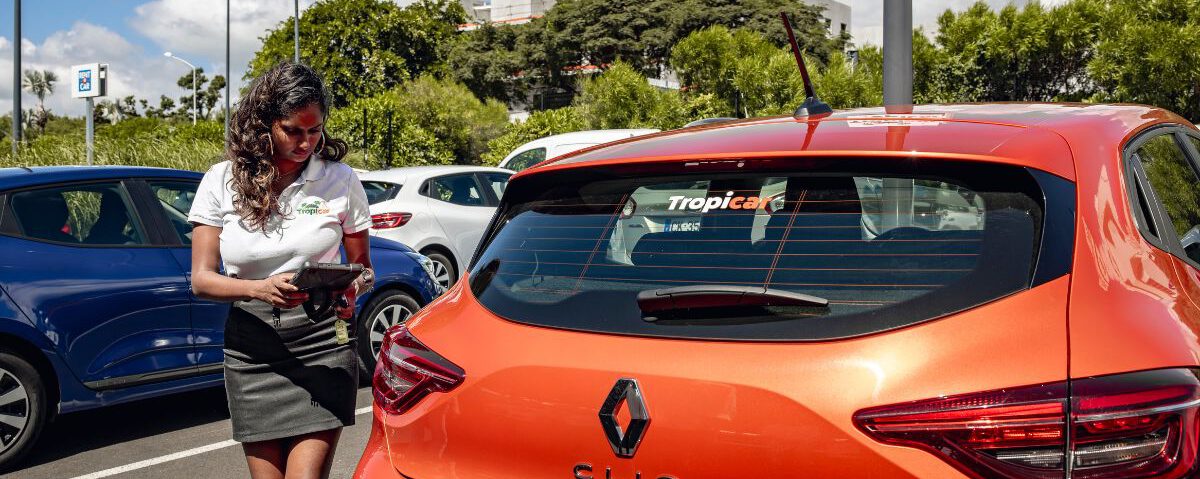 Une hôtesse Tropicar souriante et professionnelle effectue un état des lieux minutieux d'une Renault Clio sur le parking de l'aéroport Roland Garros à La Réunion